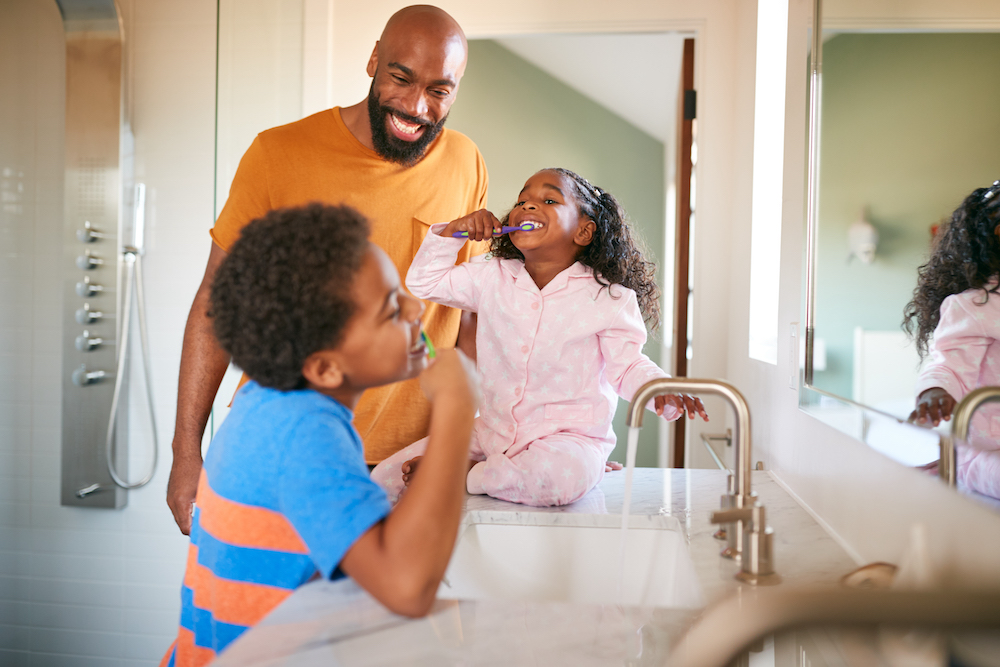 Father Helping Children To Brush Teeth In Bathroom At Home