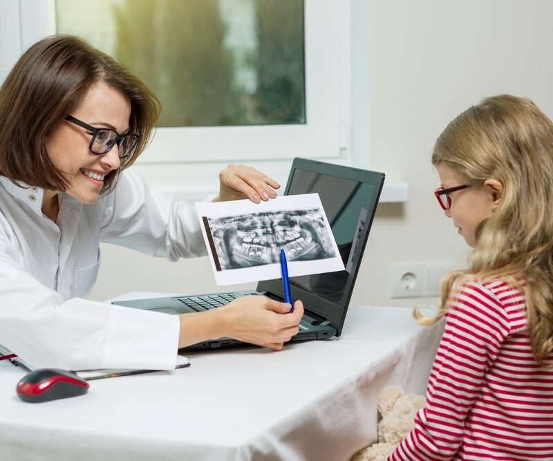 Pediatric dentist showing dental x ray to child during consultation before school appointment in clinic